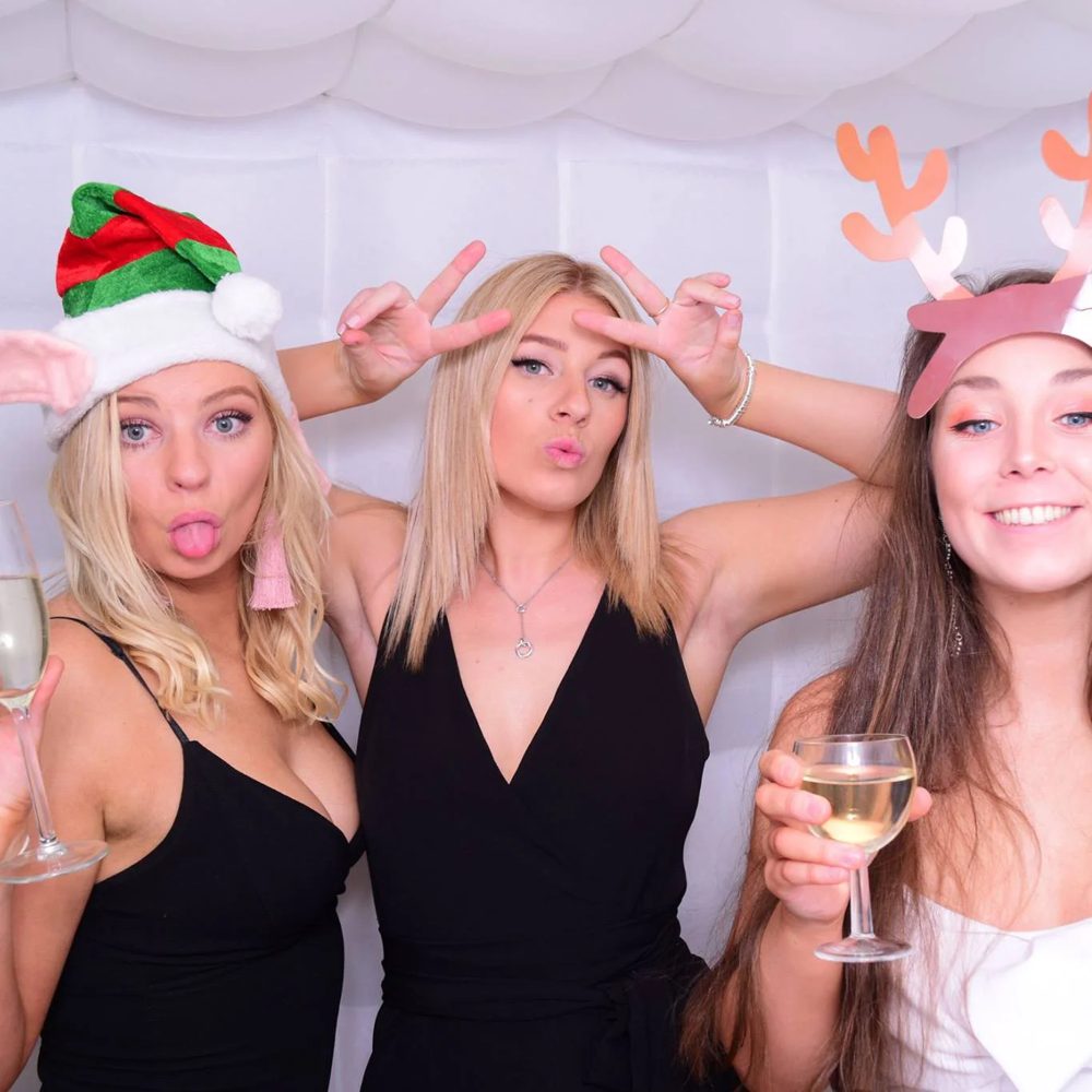 Friends in Santa hats and reindeer antlers with drinks inside the white inflatable Bubble photo booth at a Christmas party